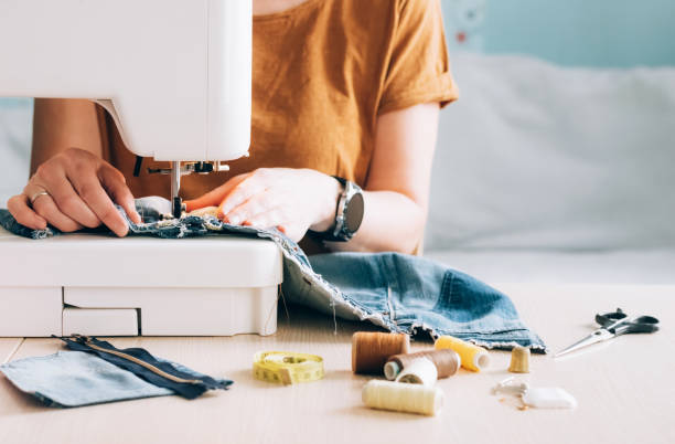 A woman tailor works at sewing machine sews reuses fabric from old denim clothes A woman tailor works at a sewing machine sews reuses fabric from old denim clothes. The concept of economical reuse of recyclable things. Homemade needlework hobby. Selective focus hole in pants stock pictures, royalty-free photos & images