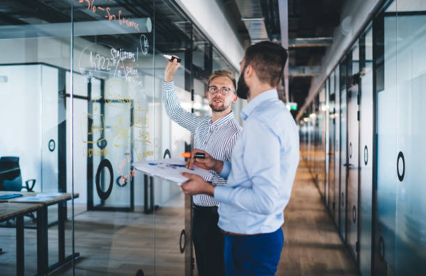 istock Confident male boss looking at his employee explaining data and information writing on glass and communicating, young 20s men business partners solving problem having productive job on meeting in office 1301209935