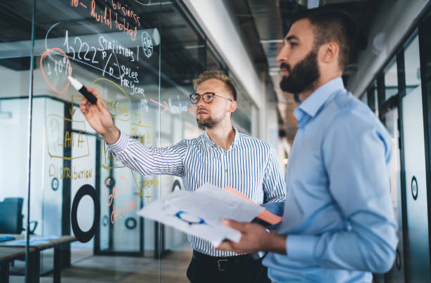 colegas de hombres casuales inteligentes que tienen cooperación productiva en ideas de compartir trabajo durante la conversación productiva en el espacio de trabajo moderno con tablero de vidrio, vendedores masculinos confiados discutiendo informe analí - economist fotografías e imágenes de stock