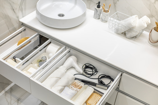 Bathroom under sink organizer drawers with neatly placed bath amenities and toiletries. Table top basin sink with dispenser of liquid soap, toothbrush in cup and group of rolled up white towels.