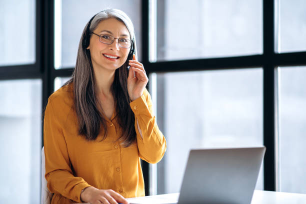 mulher ou tutor de cabelos grisalhos asiáticos simpáticos, em roupas casuais, fone de ouvido e óculos está trabalhando online, ou realizar ensino à distância, usando laptop no local de trabalho, olha para a câmera, sorri - looking at camera senior women laptop only women - fotografias e filmes do acervo