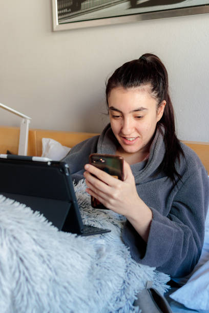 Young woman studying at home on the cam a Young Caucasian woman smiling sitting on the bed in pajamas wearing the tablet and mobile studying remotely in her room confinamiento stock pictures, royalty-free photos & images