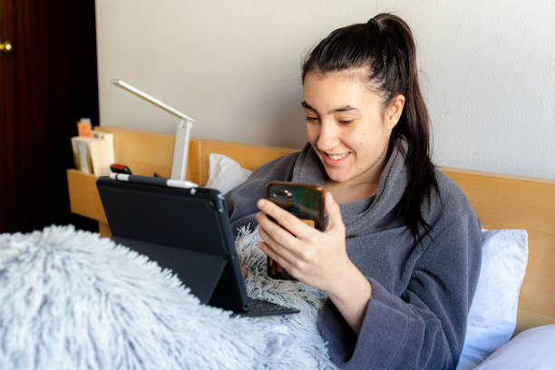 Young woman studying at home on the cam a Young Caucasian woman smiling sitting on the bed in pajamas wearing the tablet and mobile studying remotely in her room confinamiento stock pictures, royalty-free photos & images