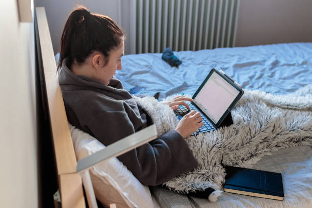 Young woman writing with her tablet in bed Young Caucasian woman sitting on the bed in her room in her pajamas covered with a blanket using a tablet studying at home. Has a notebook above the bed confinamiento stock pictures, royalty-free photos & images