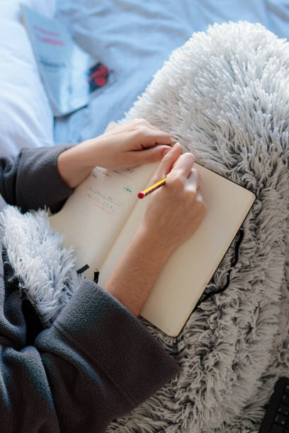 Woman's close-up writing on the bed Young Caucasian woman writing in a notebook with a pencil on top of her skirt covered with a blanket on top of her room bed in the morning. Study at home confinamiento stock pictures, royalty-free photos & images