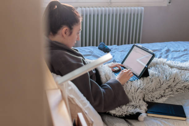 Young woman writing with her tablet in bed Young Caucasian woman sitting on the bed in her room in her pajamas covered with a blanket using a tablet studying at home. Has a notebook above the bed confinamiento stock pictures, royalty-free photos & images