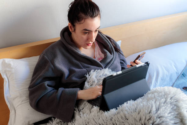 Young woman using tablet and mobile while studying on the bed Young Caucasian woman sitting in bed in pajamas wearing the tablet and consulting something on the mobile phone studying remotely in her room above the bed confinamiento stock pictures, royalty-free photos & images