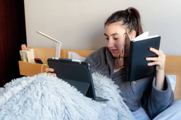 Young Caucasian woman studying in bed Young Caucasian woman sitting on the bed covered with a blanket in pajama clothes holding a notebook with one hand and with a tablet in her skirt styling from home in her room in the morning. confinamiento stock pictures, royalty-free photos & images