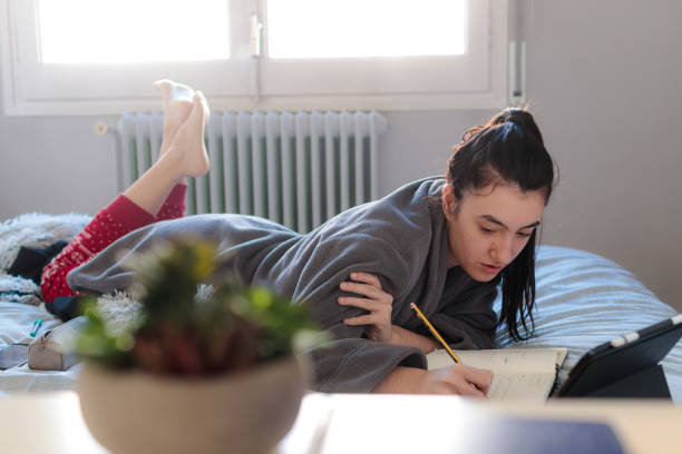 Young woman sceping in a notebook over the bed Young Caucasian woman writing in a notebook with a pencil stretched out on top of the bed in pajamas next to a tablet in the morning. Study at home confinamiento stock pictures, royalty-free photos & images