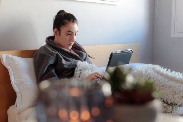 Young woman writing with her tablet in bed Young Caucasian woman sitting on the bed in her room in her pajamas covered with a blanket using a tablet studying at home. confinamiento stock pictures, royalty-free photos & images