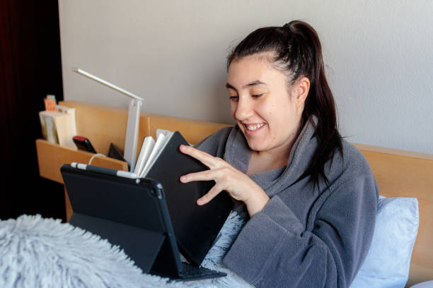 Young woman studying in bed smiling Young Caucasian woman sitting on the bed covered with a blanket in pajama clothes holding a notebook with one hand and with a tablet in her skirt styling from home in her room in the morning. confinamiento stock pictures, royalty-free photos & images