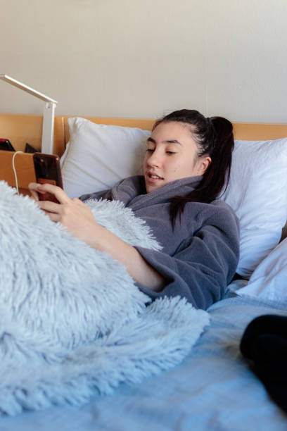 Young woman looked at her cell phone in bed Young Caucasian woman smiling looking at the mobile phone stretched out on the bed in pajama clothes covered by a blanket confinamiento stock pictures, royalty-free photos & images