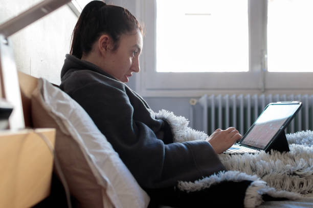 Young woman writing with her tablet in bed Young Caucasian woman sitting on the bed in her room in pajama clothes covered with a blanket using a tablet studying at home in the morning. Study at home. confinamiento stock pictures, royalty-free photos & images