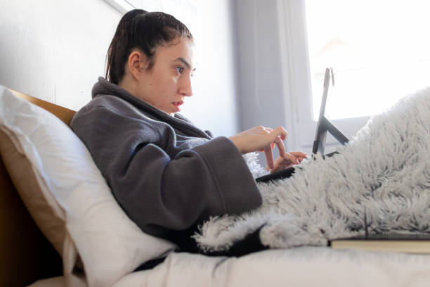 Young woman writing with her tablet in bed Young Caucasian woman sitting on the bed in her room in her pajamas covered with a blanket using a tablet studying at home. confinamiento stock pictures, royalty-free photos & images