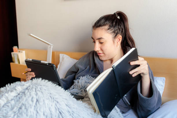 Young Caucasian woman studying in bed Young Caucasian woman sitting on the bed covered with a blanket in pajama clothes holding a notebook with one hand and with a tablet in her skirt styling from home in her room in the morning. confinamiento stock pictures, royalty-free photos & images