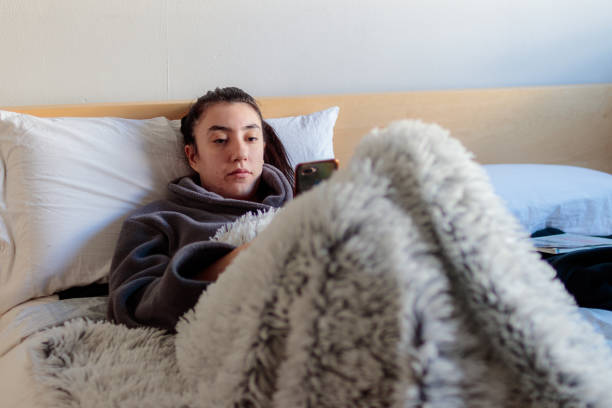 Young woman using mobile phone in bed A young Caucasian woman in pajama clothes is sitting on the bed watching and using her smiling mobile phone covered with a blanket in her room in the morning. confinamiento stock pictures, royalty-free photos & images