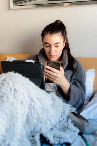 Young woman in bed looking at her cell phone while she's studying A young Caucasian woman sitting on her bedroom bed in pajama clothes covered with a blanket next to a tablet. Study at home confinamiento stock pictures, royalty-free photos & images