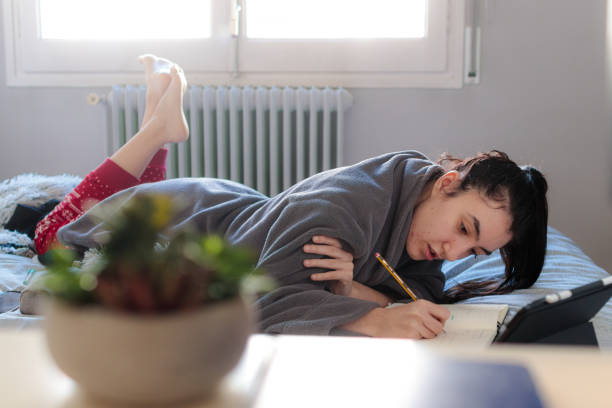 Young woman sceping in a notebook over the bed Young Caucasian woman writing in a notebook with a pencil stretched out on top of the bed in pajamas next to a tablet in the morning. Study at home confinamiento stock pictures, royalty-free photos & images