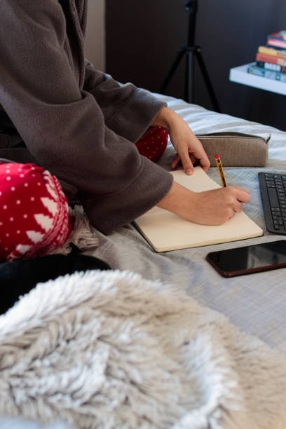 Young woman sceping in a notebook over the bed Young Caucasian woman writing in a notebook with a pencil stretched out on top of the bed in pajamas next to a tablet in the morning. Study at home confinamiento stock pictures, royalty-free photos & images