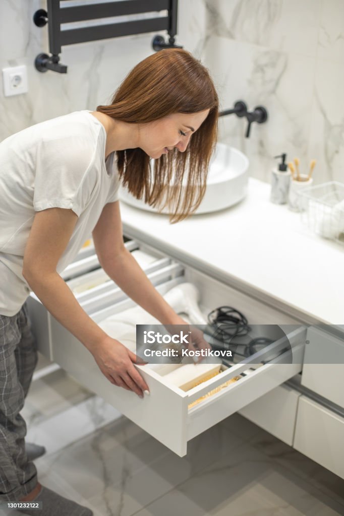 Happy young woman standing near dressing table and taking out a beauty cosmetic from drawer Happy young woman standing in front of open drawers of dressing table and taking out toiletries from drawer. Concept of saving space and organizing vanity table. Selective focus. Bathroom Stock Photo