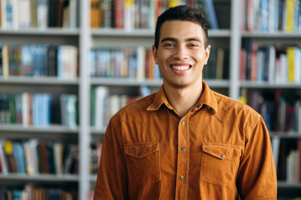 verticale de l’homme hispanique réussi d’affaires regarde directement à l’appareil-photo, sourire. pigiste ou étudiant mâle heureux restant dans le bureau moderne ou la bibliothèque d’université - latino américain photos et images de collection