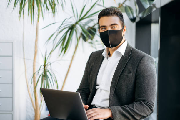istock Confident man in formal wear wearing a protective black mask on his face works remotely using a laptop while sitting indoor and looking directly into camera 1301235044