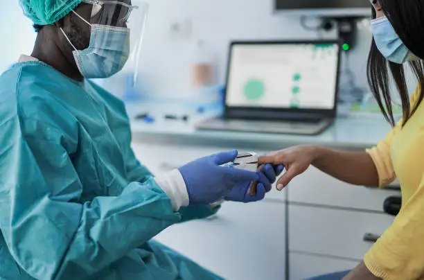 African male doctor using pulse oximeter on young woman patient finger while wearing surgical face masks for coronavirus outbreak - Measuring oxygen saturation