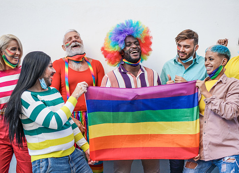 Multiracial gay people having fun dancing at lgbt pride parade while wearing protective face masks under chin