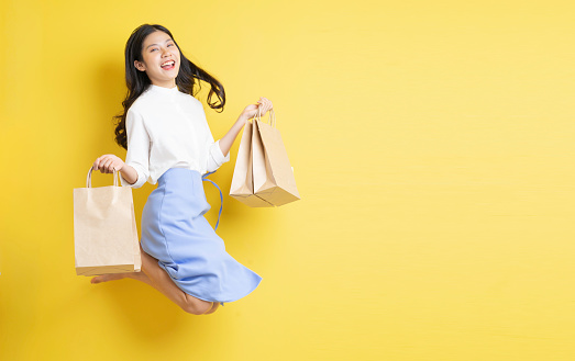 Young girl holding shopping bag with cheerful expression jumping up on yellow background