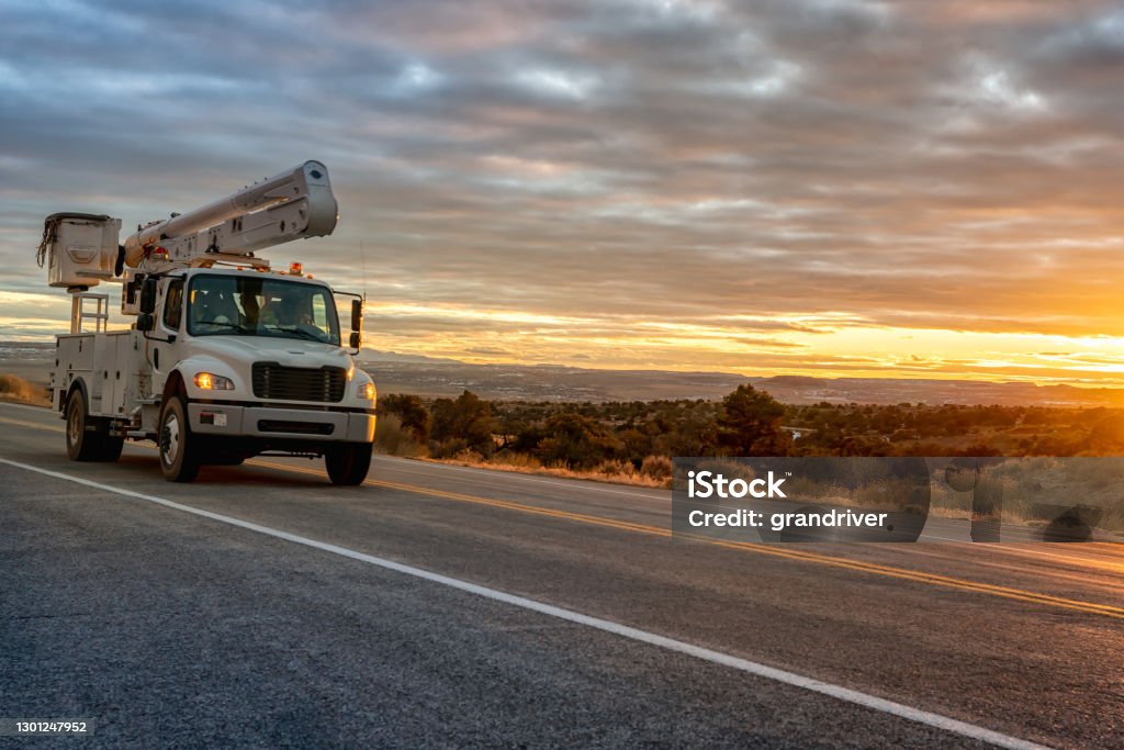 A Bucket Or Aerial Platform, Boom Truck Driving Through The Desert Of Utah, Near Moab, Wilson Arch, With A Dramatic Sunset To The West A Bucket Or Boom Truck Driving Through The Desert Of Utah, Near Moab, Wilson Arch, With A Dramatic Sunset To The West Truck Stock Photo