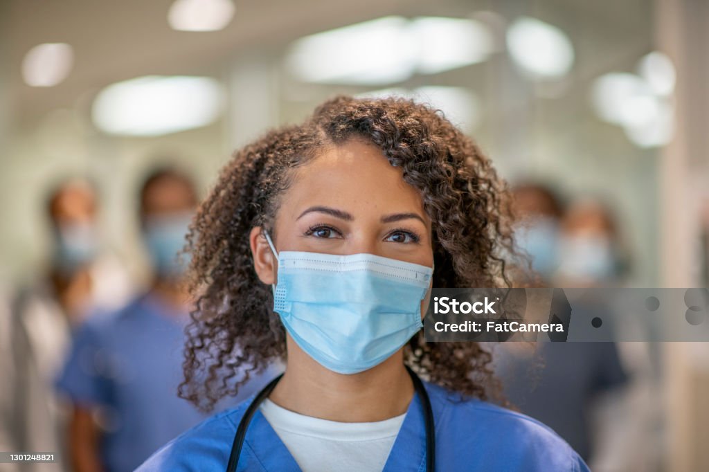 Beautiful female doctor smiling behind her mask A female doctor of African descent is wearing blue medical scrubs and a face mask stands in front of her colleagues at the hospital. She is smiling behind her mask. Nurse Stock Photo