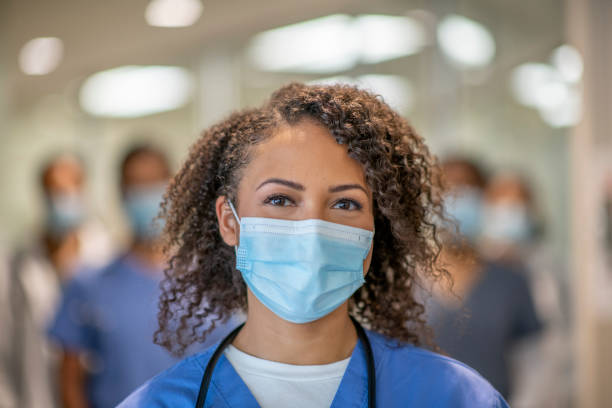 Beautiful female doctor smiling behind her mask A female doctor of African descent is wearing blue medical scrubs and a face mask stands in front of her colleagues at the hospital. She is smiling behind her mask. protective face mask stock pictures, royalty-free photos & images