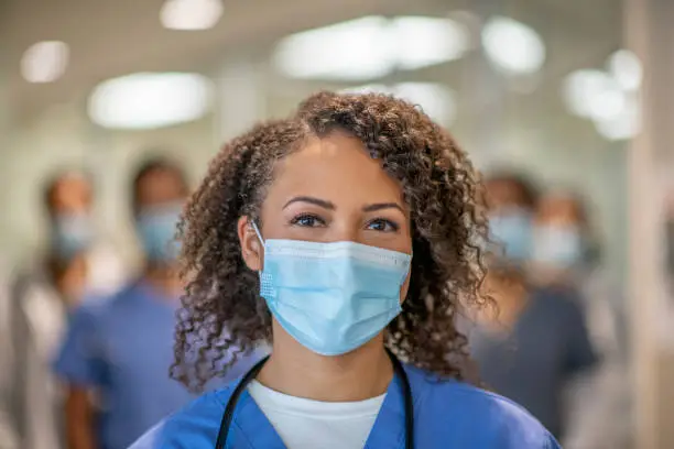 A female doctor of African descent is wearing blue medical scrubs and a face mask stands in front of her colleagues at the hospital. She is smiling behind her mask.