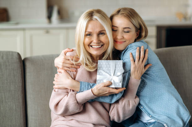 überraschung für muttertag oder geburtstag. liebevolle junge erwachsene tochter, die ihrer geliebten kaukasischen mutter mittleren alters ein geschenk schenkt, die auf dem sofa im wohnzimmer sitzt - living room people joy happiness stock-fotos und bilder