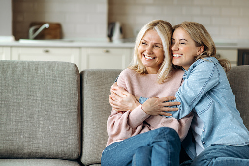 Portrait of caucasian mother and daughter. Mature attractive mother and her young adult happy daughter, are sitting at home on the couch, embracing, smiling. Family values