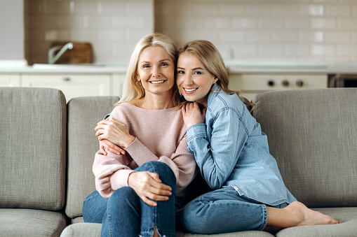 Portrait of caucasian mother and daughter. Mature attractive mother and her young adult happy daughter, are sitting at home on the couch, embracing, looking at the camera and smiling. Family values