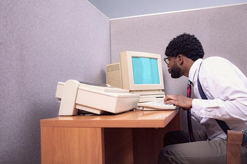 A vintage African American business man at the office works at an old computer at his cubicle desk.  1980's - 1990's fashion style.