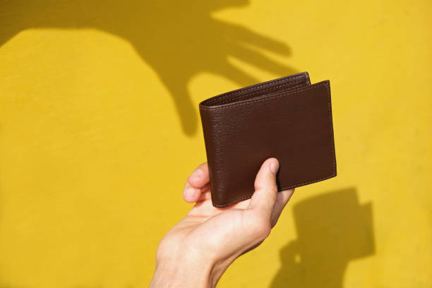 Shadow of robbers hand on yellow wall trying to steal leather wallet from persons hand. Pickpocket silhouette. Concept of financial crime, tax burden, unexpected expenses. Shadow of robbers hand on yellow wall trying to steal leather wallet from persons hand. Pickpocket silhouette. Concept of financial crime, tax burden, unexpected expenses. pickpocketing stock pictures, royalty-free photos & images