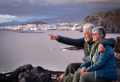 Beautiful embraced senior couple sitting on the cliff on a winter day enjoying the sunset over the sea. New young people enjoy freedom and retirement. Cloudy sky, coast and mountain in the background
