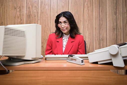 A vintage Filipino business woman at the office works at an old computer at her desk.  1980's - 1990's fashion style.  Wood paneling on wall in the background.