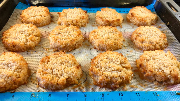 Close-up image rows of freshly baked coconut macaroon cakes on oven baking tray lined with silicone baking sheet, focus on foreground Stock photo showing oven baking tray lined with silicone baking sheet containing freshly baked coconut macaroons cakes, made with desiccated coconut mixed with egg whites, sugar, butter and vanilla flavour. Coconut biscuit stock pictures, royalty-free photos & images