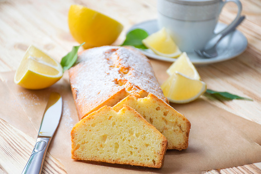 Moist lemon pound cake on parchment on rustic wooden background with slices of lemon, knife and cup of tea on plate. Delicious breakfast, traditional English tea time. Reciepe of lemon pie loaf.