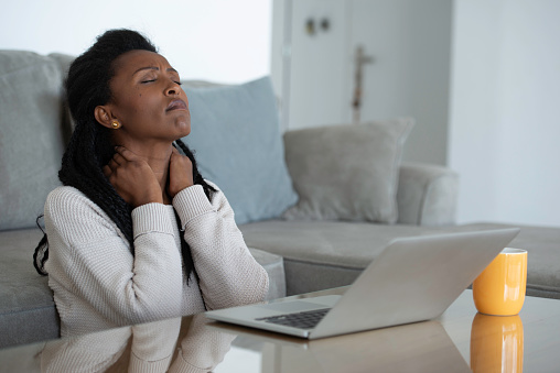 Young African woman sitting in front of the computer, working from home online, drinking coffee, looks tired and nervous. The woman trying to reduce neck tension.