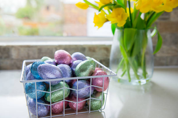 metallkorb mit schoko-ostereiern in farbfolie und strauß gelber tulpen und narzissen blühen auf weißem küchentisch in der nähe des fensters. festliche frühlingskomposition. selektiver fokus, kopierraum - daffodil easter egg hunt easter easter egg stock-fotos und bilder