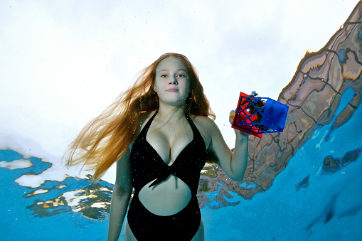Portrait of a young girl with long hair swimming underwater in a pool with a toy in her hand in a black swimsuit on a sunny day on a blue background. The concept. Wide angle. Bottom view