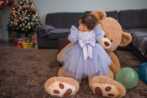 Cute smiling baby girl in dress playing with her teddy bear in living room