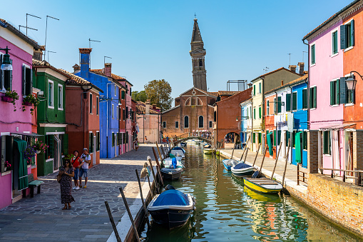Burano, Italy, Sep. 2020 – A colorful canal in Burano with the leaning bell tower of San Martino church in the background