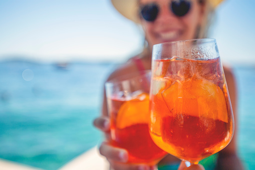 Couple toasting with a spritz cocktail on a deck beside the ocean. The women is wearing a bikini sunglasses and hat and is smiling. Turquoise ocean.  Selective focus with Copy space