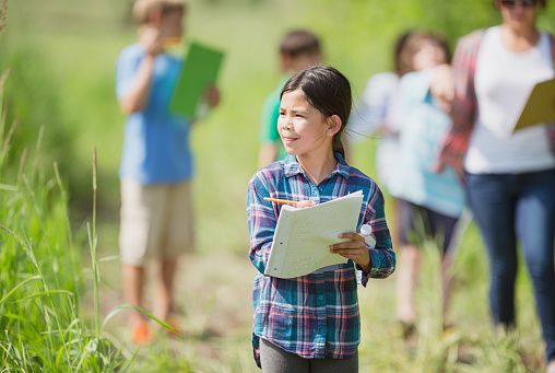 An elementary school girl of Filipino ethnicity is outdoors with her class exploring nature. She is busy writing on her notepad while walking with the group.