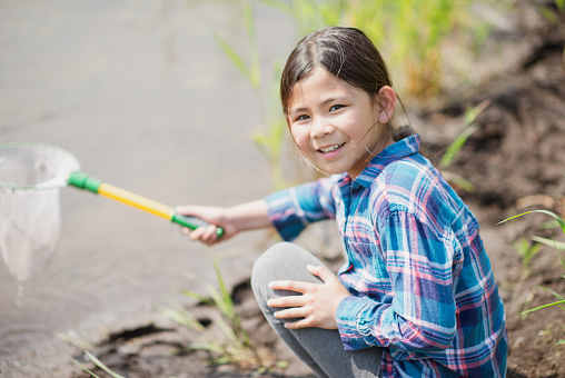 An adorable elementary school girl of Filipino ethnicity is outdoors on a warm summer day exploring nature. She is holding a butterfly next to to muddy waters. She is smiling at the camera.